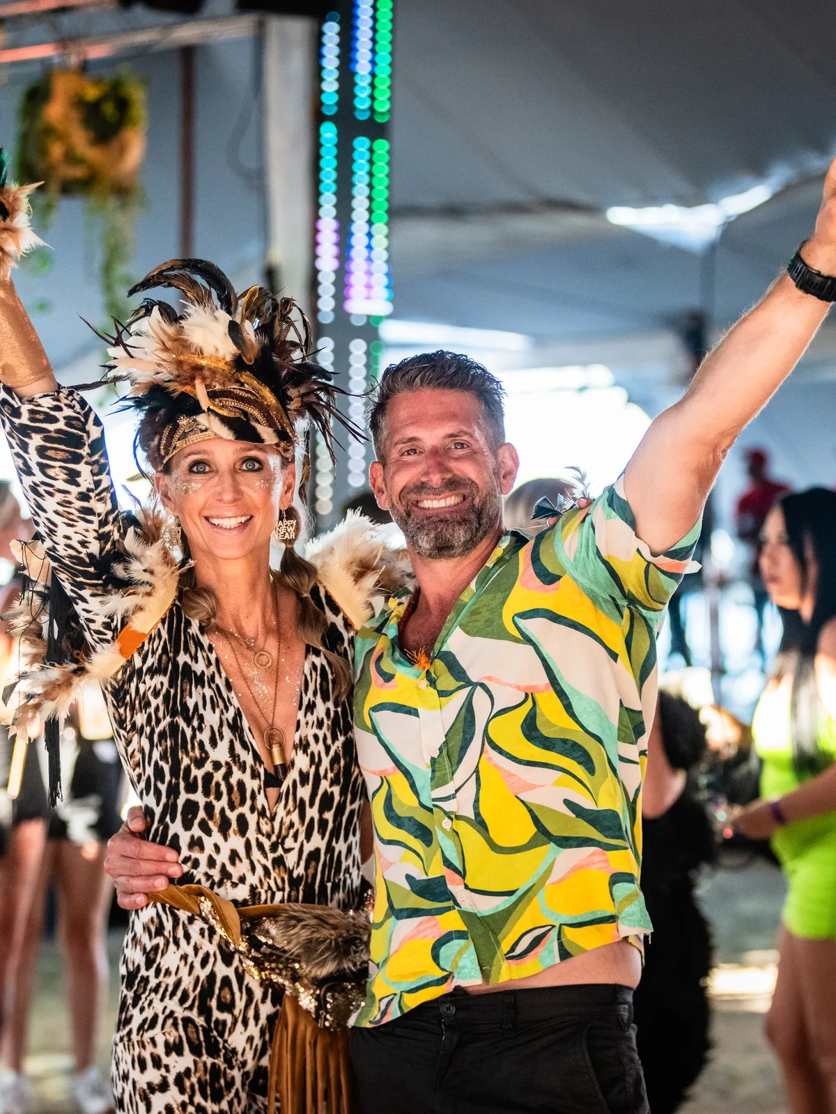 Two guests in costume, a feathered headdress and a leaf-print shirt, arms raised